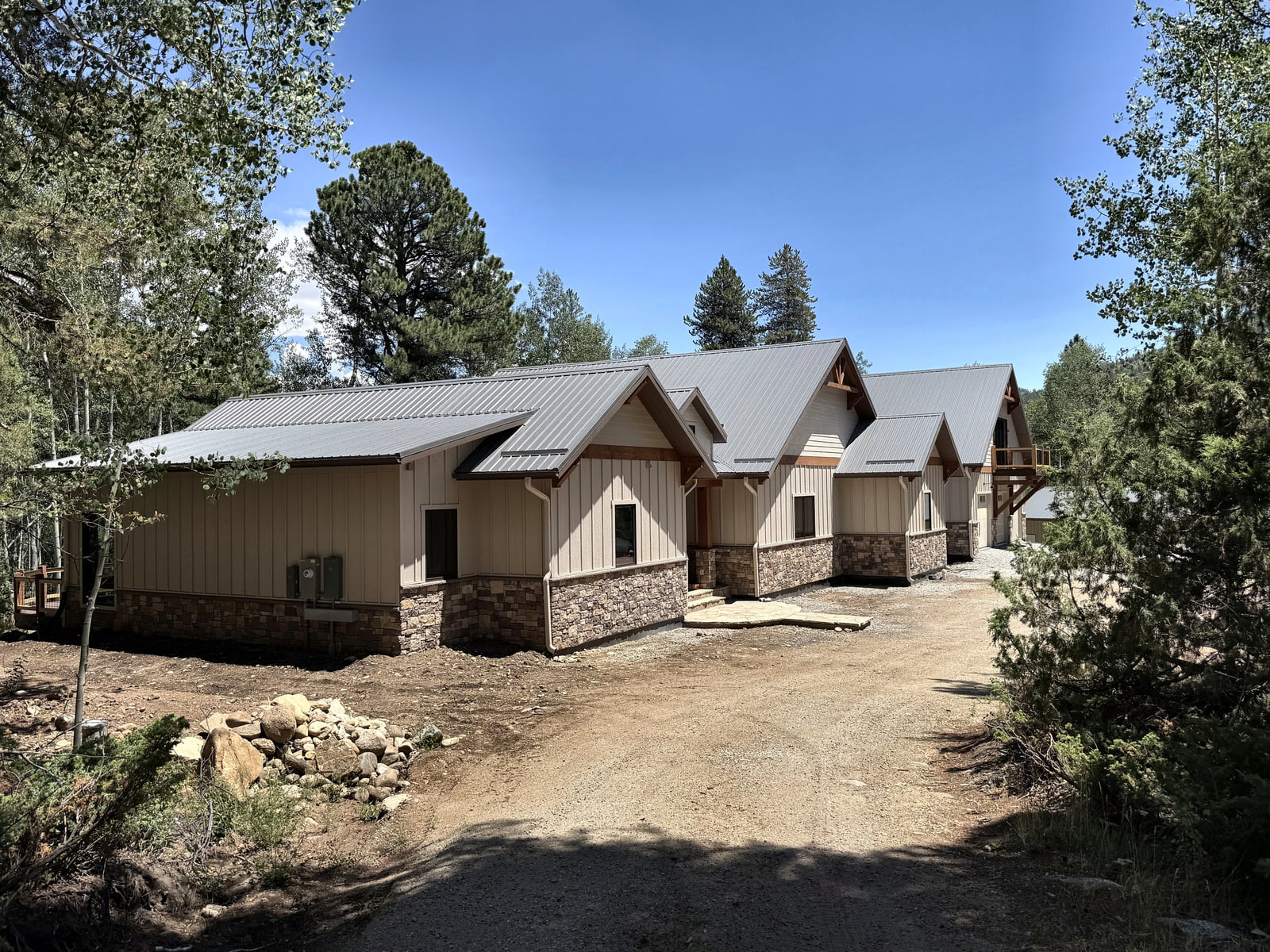 Completed photograph: Front approach to a custom mountain residence with metal gable roofs, stone base, and natural wood accents.