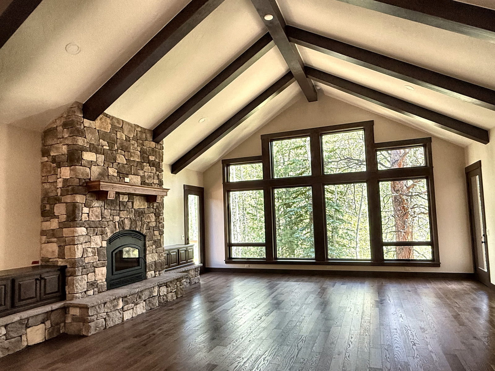 Vaulted great room with exposed dark beams, stone fireplace, wood floor, and large windows overlooking the forest.